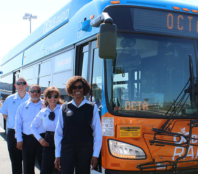 A group of smiling OC Bus drivers stand in a row in front of an OC Bus