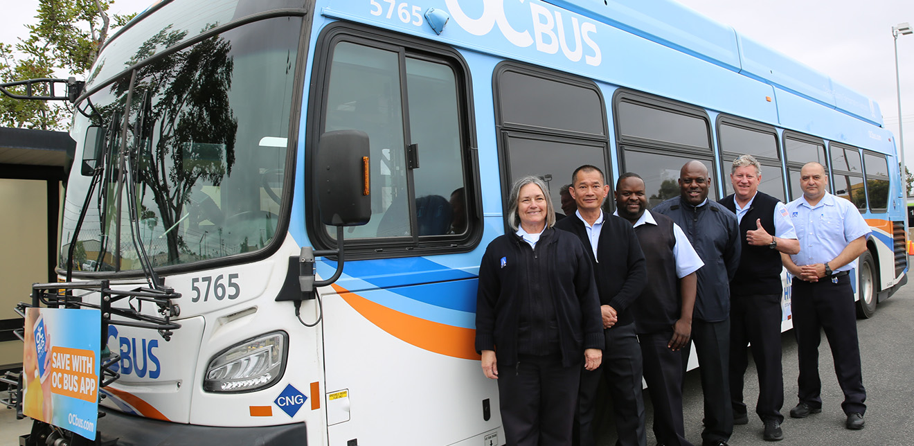 A group of smiling OC Bus drivers stand in a row in front of an OC Bus