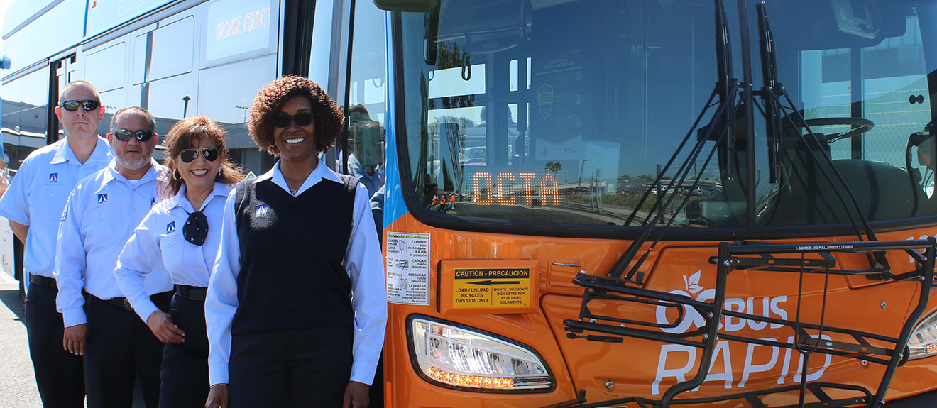 A group of smiling OC Bus drivers stand in a row in front of an OC Bus