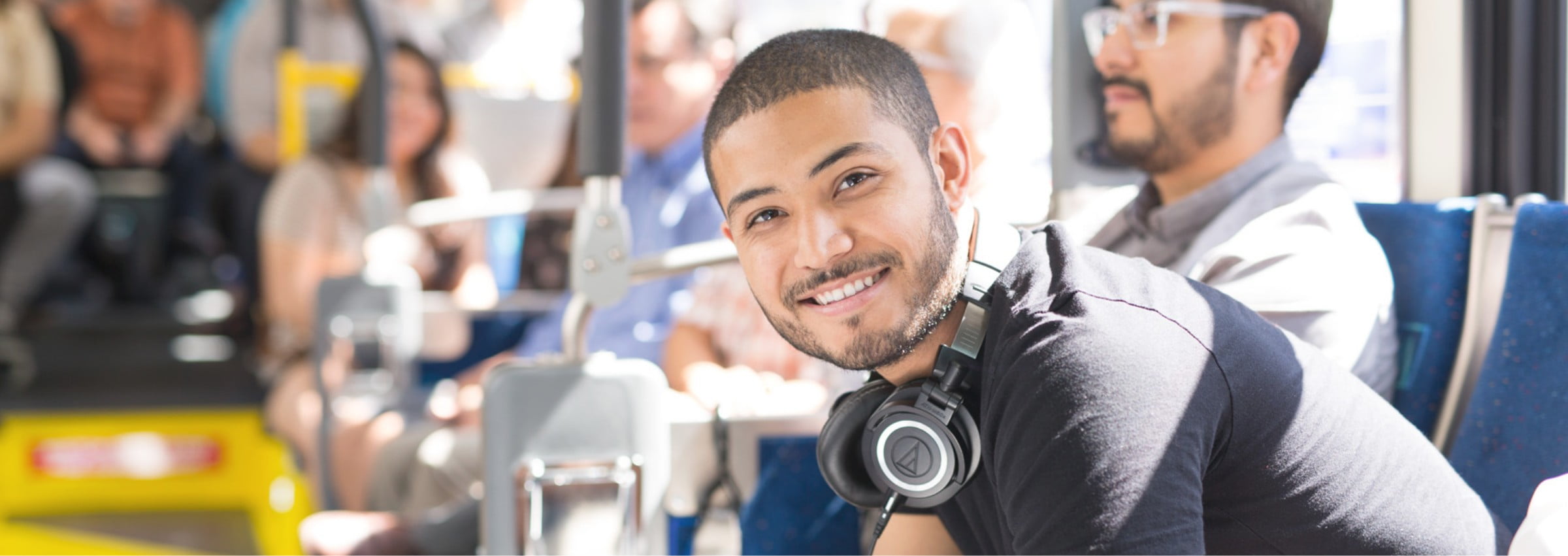 A young male rider smiles on the bus while wearing headphones around his neck